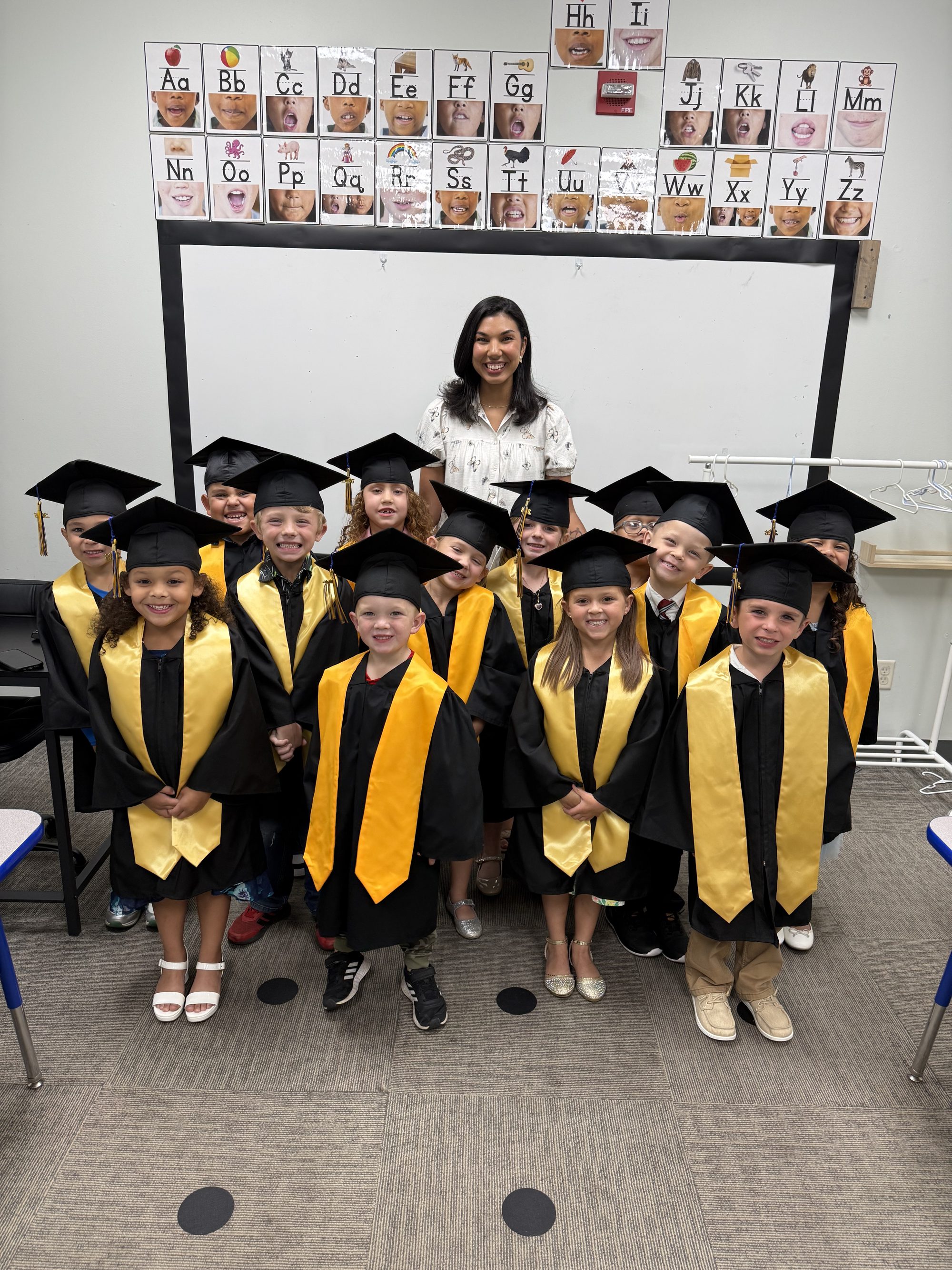 Kindergarten graduates in black and gold gowns celebrating with their teacher